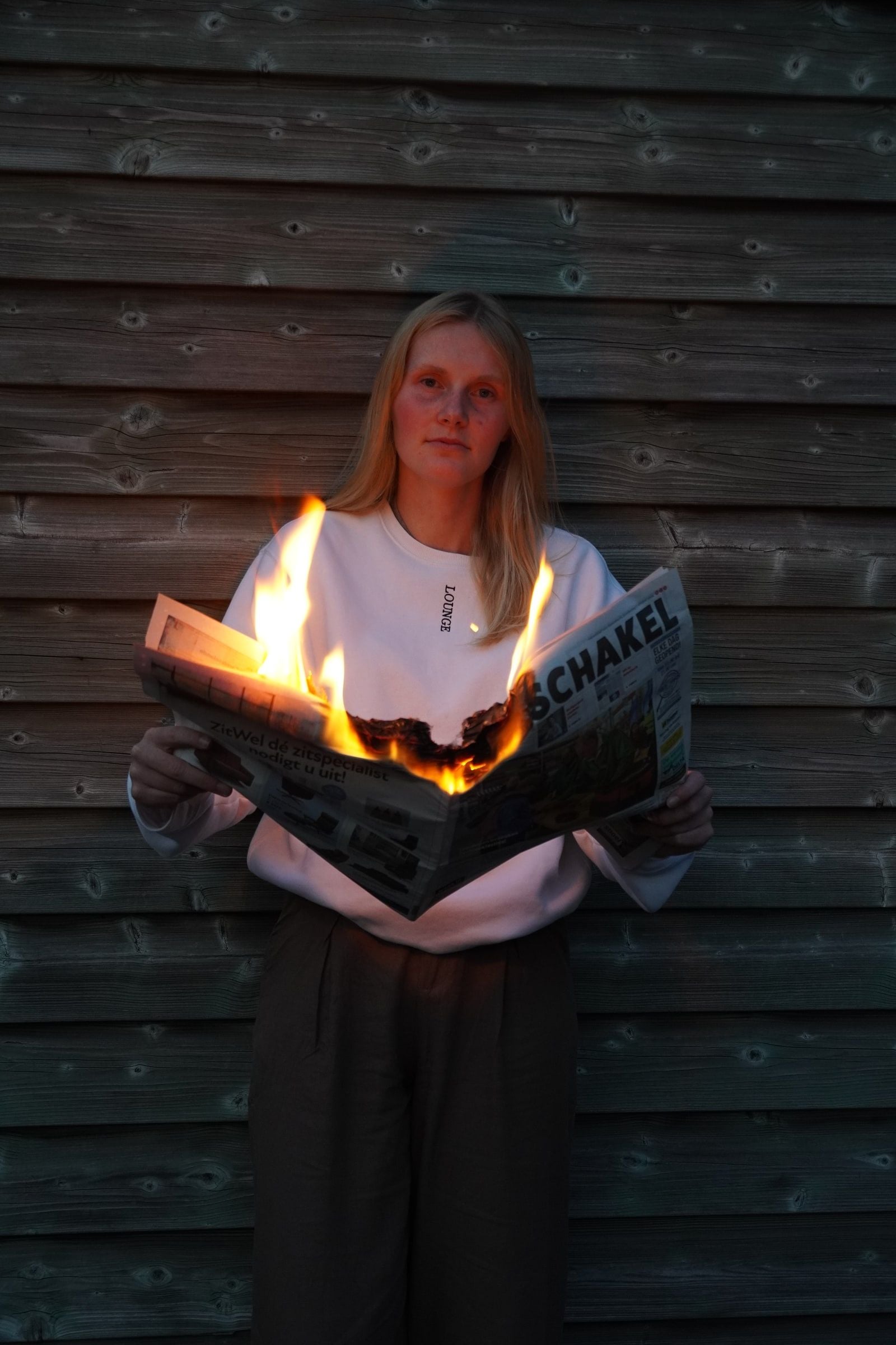 Persoon met lang blond haar en witte sweater staat voor een houten schuurwand en houdt een opengeslagen krant vast die in het midden in brand staat; het vuur verlicht de krant en de onderkant van de sweater oranje, terwijl de rest van het beeld in koel schemerlicht blijft