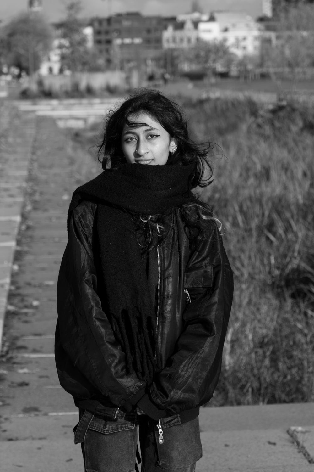 Zwart-wit portret van een persoon met donker krullend haar in een bomberjack en dikke gebreide sjaal, staand op een pad met riet en vage stadsbebouwing op de achtergrond