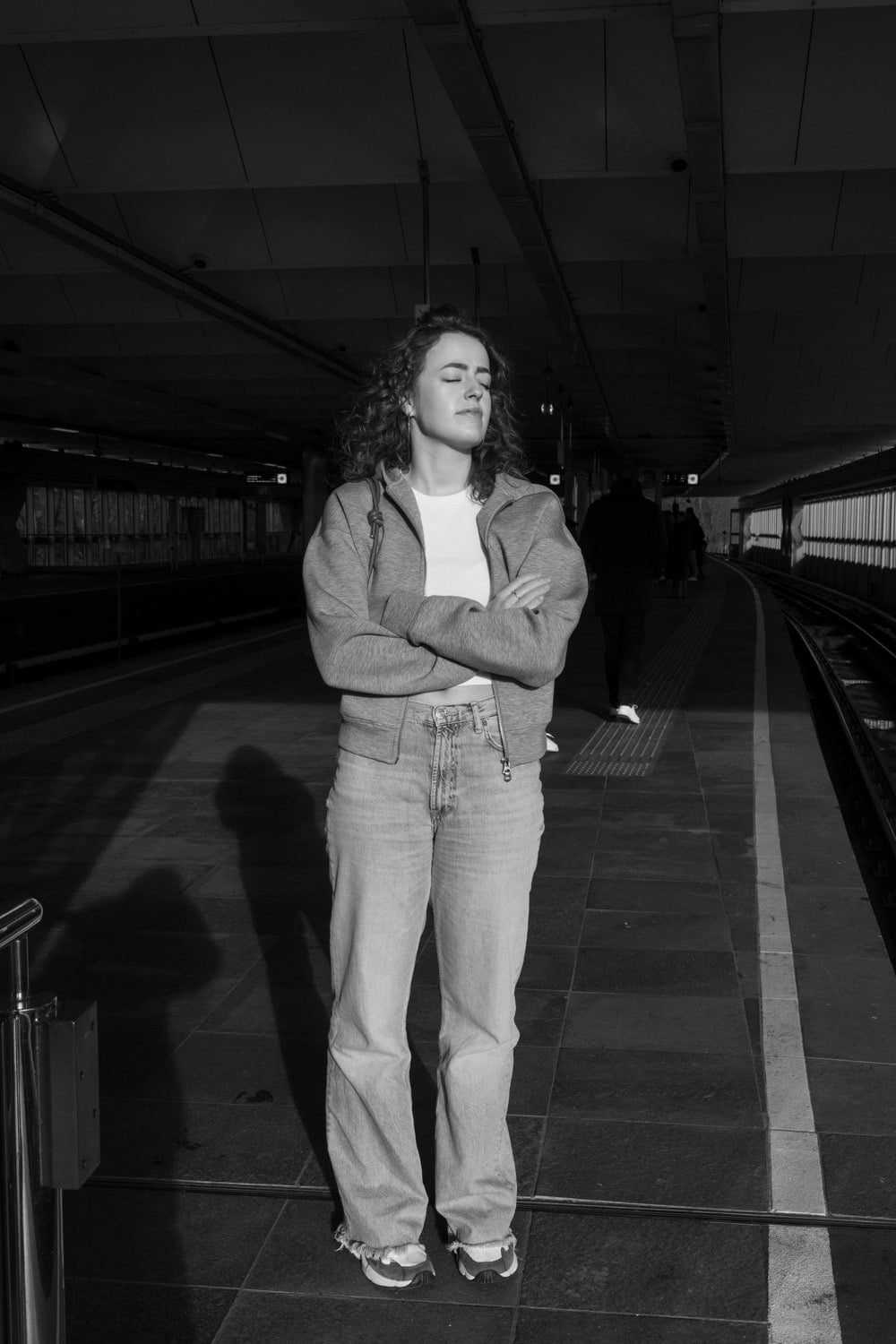 Vrouw met krullend haar staat met gesloten ogen en gekruiste armen in fel zonlicht op een overdekt metroperron; haar schaduw valt lang over de tegelvloer en achter haar loopt een donkere figuur weg over het perron. (cover)