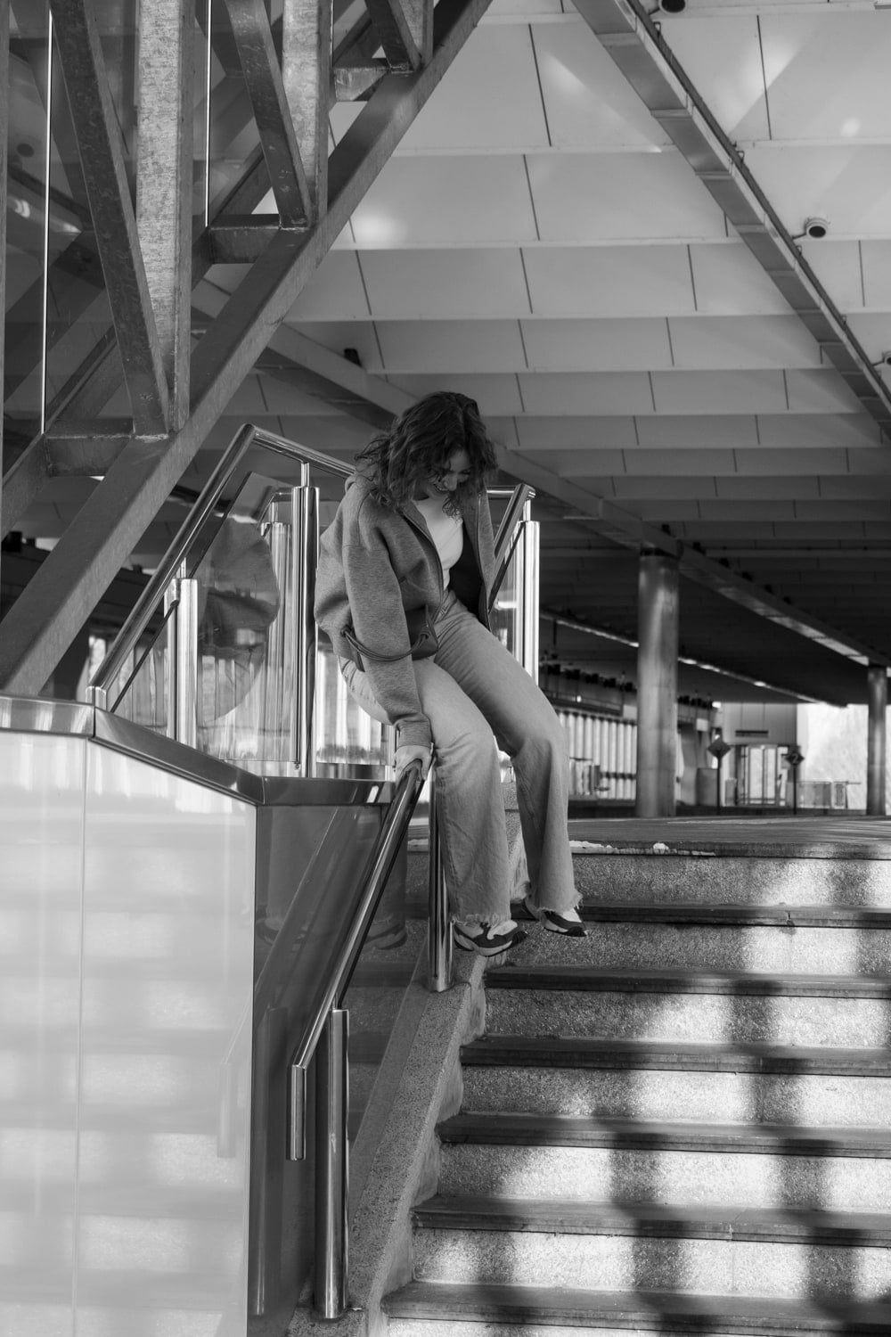 Vrouw in wollen vest en wijde jeans zit voorovergebogen op de leuning van een brede stationstrap, onder een stalen overkapping waar zonlicht in strepen over de treden valt.