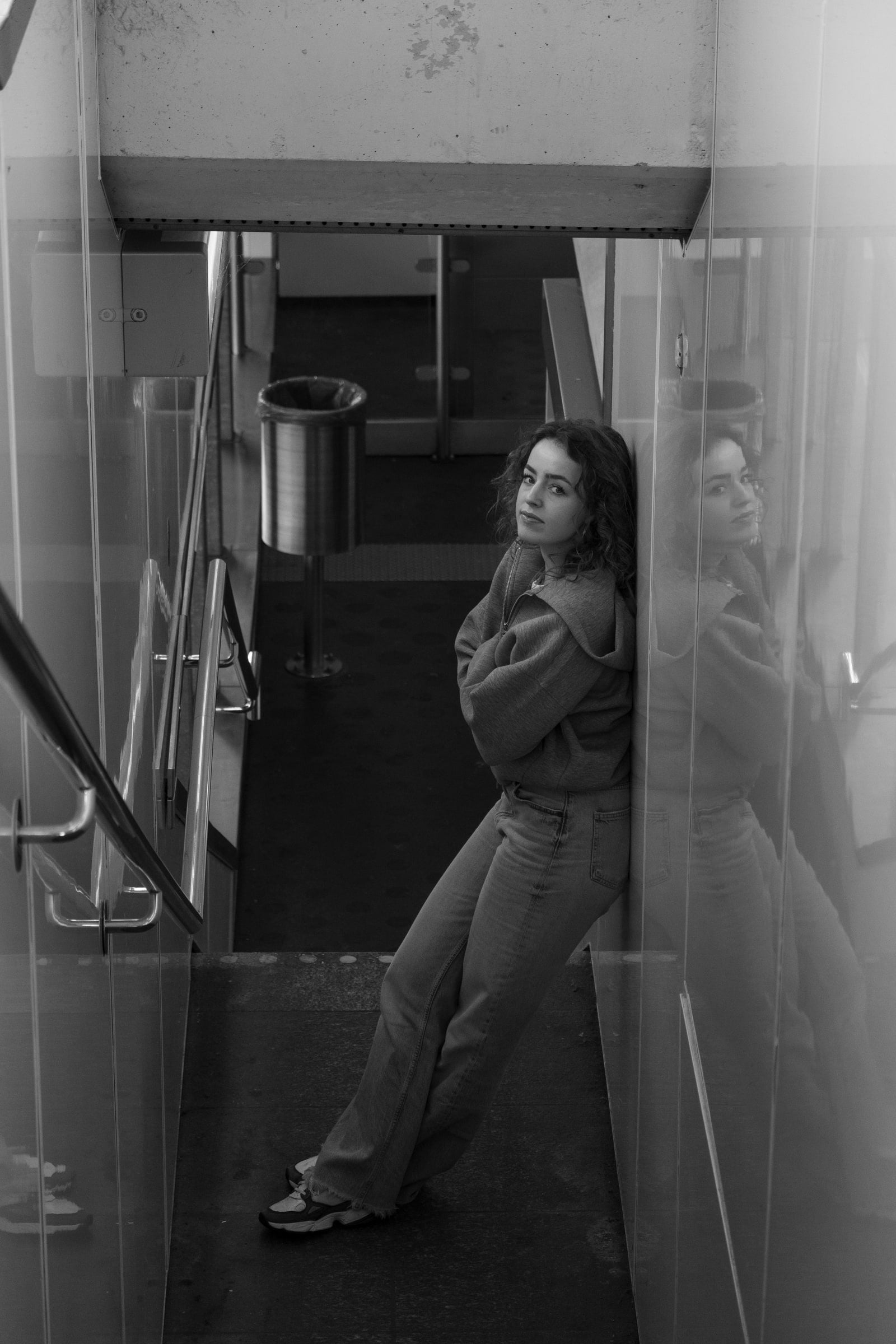Vrouw leunt met gekruiste armen tegen een glazen wand in een stationstrappenhuis; haar spiegelbeeld verschijnt naast haar in het glas, met een afvalbak en trapleuningen op de achtergrond.