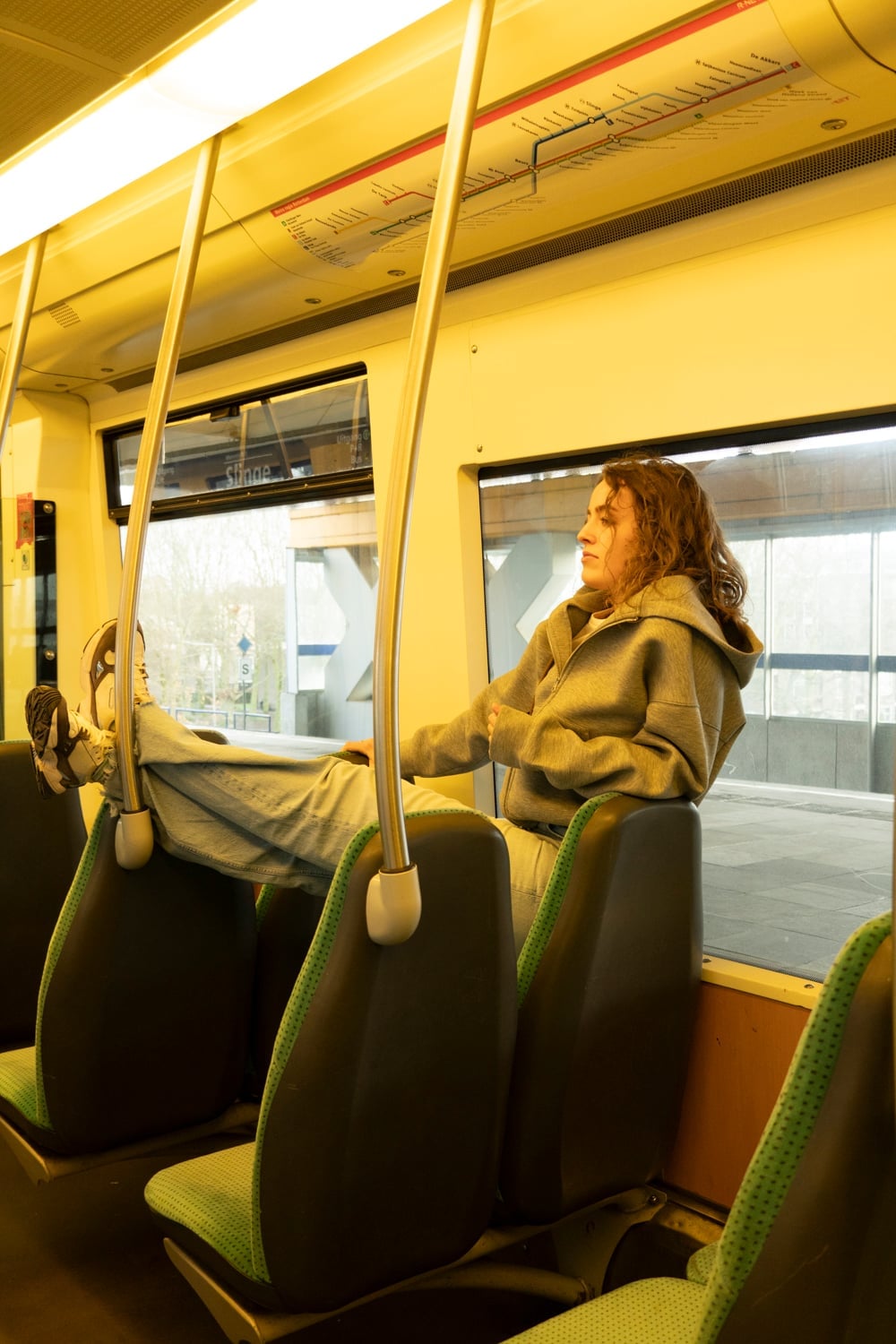 Vrouw zit achterover in een treinstoel met haar benen over de rugleuning van de bank ervoor en kijkt uit het raam naar het station dat voorbijglijdt.