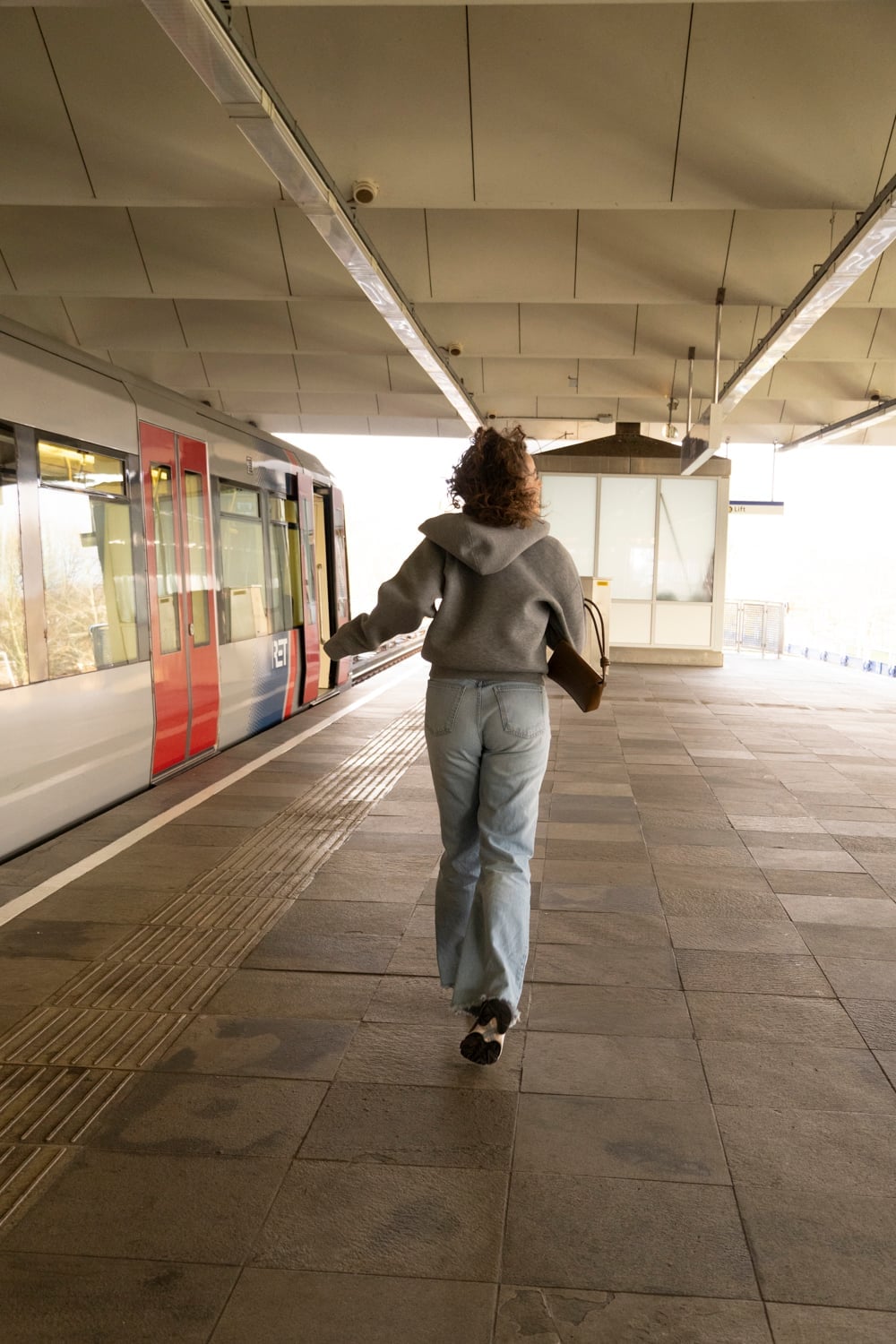 Vrouw loopt van de camera weg over een leeg perron onder een lange overkapping, met links een metro die wegrijdt.