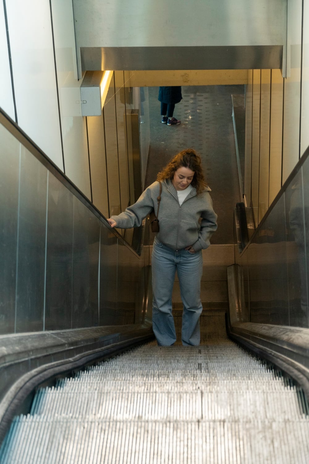 Vrouw staat bovenaan een roltrap met haar hand op de leuning en kijkt omlaag naar de treden, in een gang van geborsteld staal.