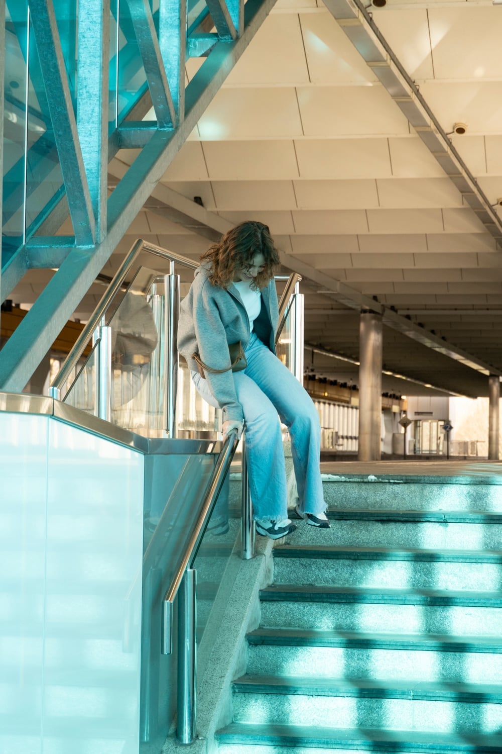 Vrouw zit voorovergebogen op de leuning van een brede stationstrap onder een blauwe stalen constructie, met zonlicht in strepen over de treden.