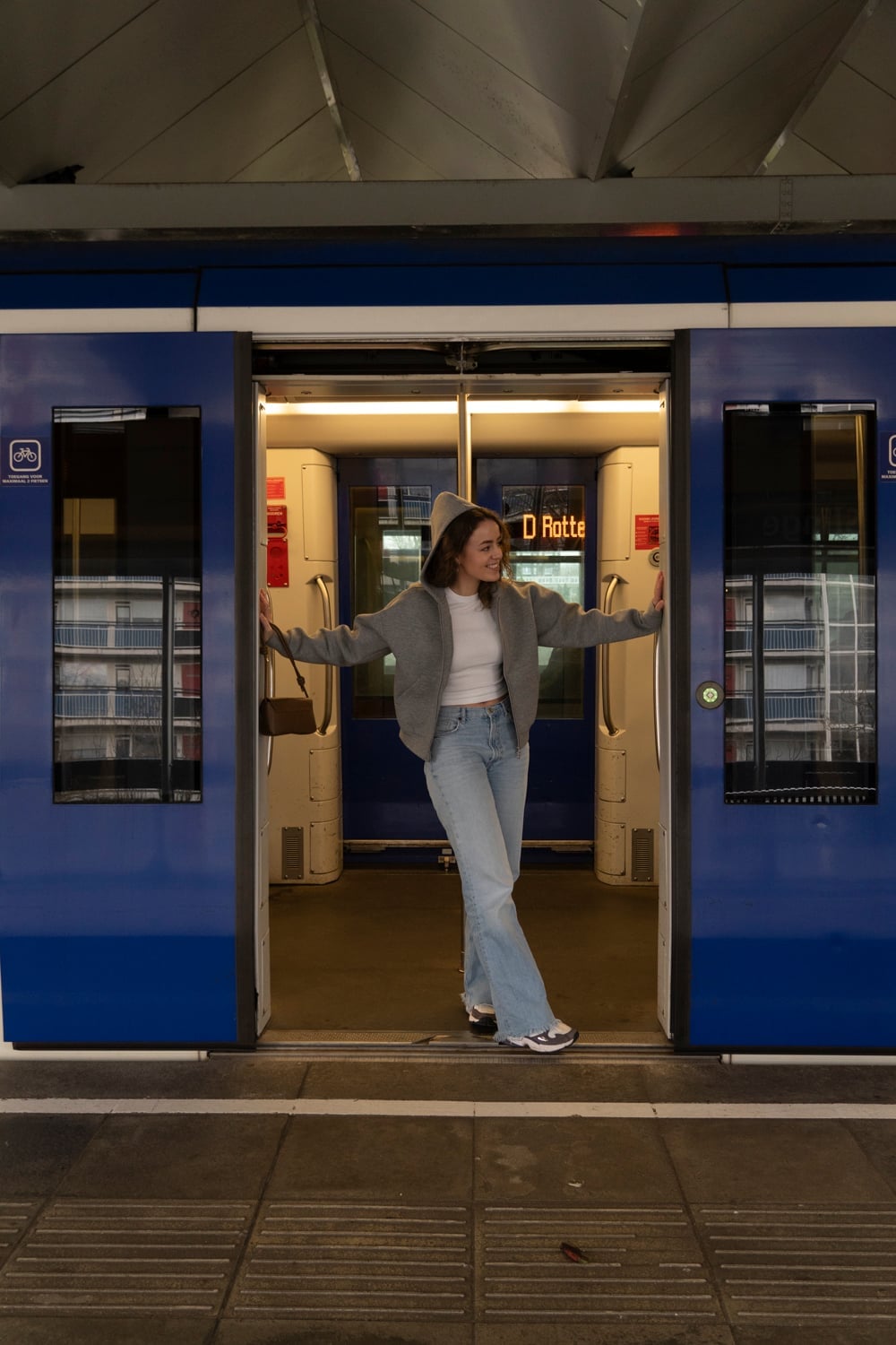 Vrouw staat met gespreide armen tussen de openstaande deuren van een blauwe trein, met de verlichte coupé achter haar.