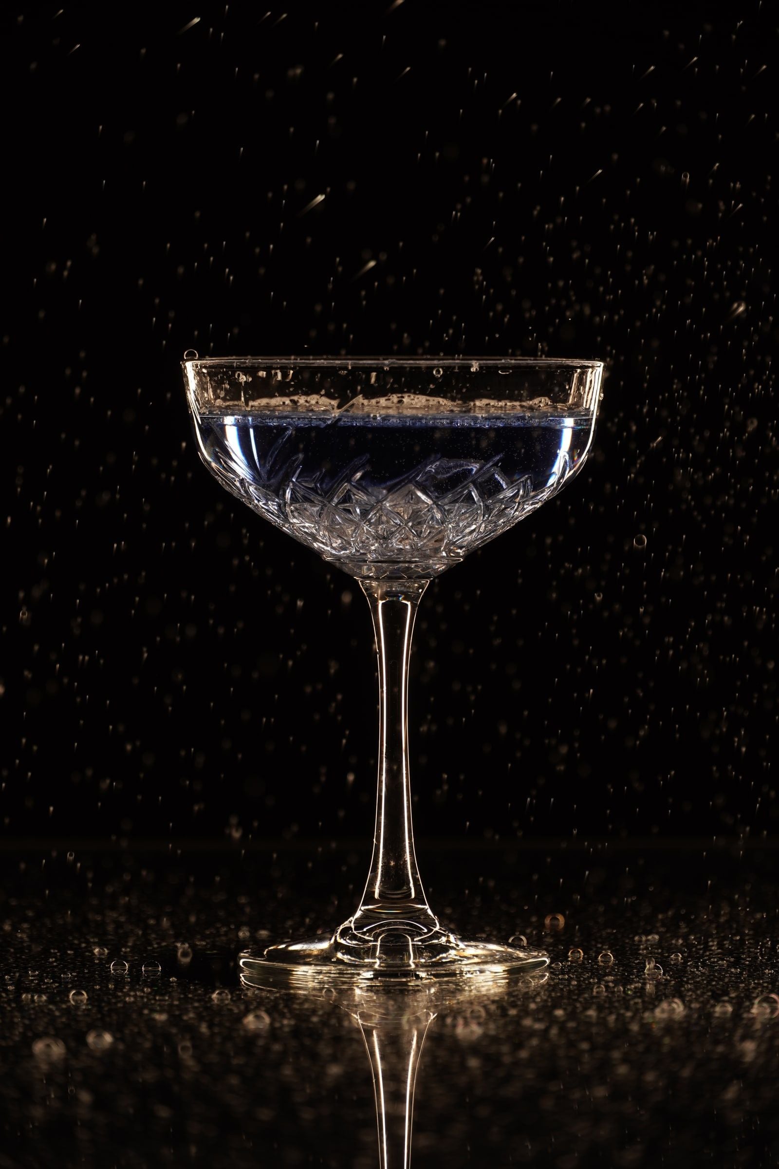 Geslepen coupeglas gevuld met blauwachtige vloeistof op een zwarte, natte ondergrond, met rondom bevroren waterdruppels in de lucht en warm randlicht langs de steel en de rand