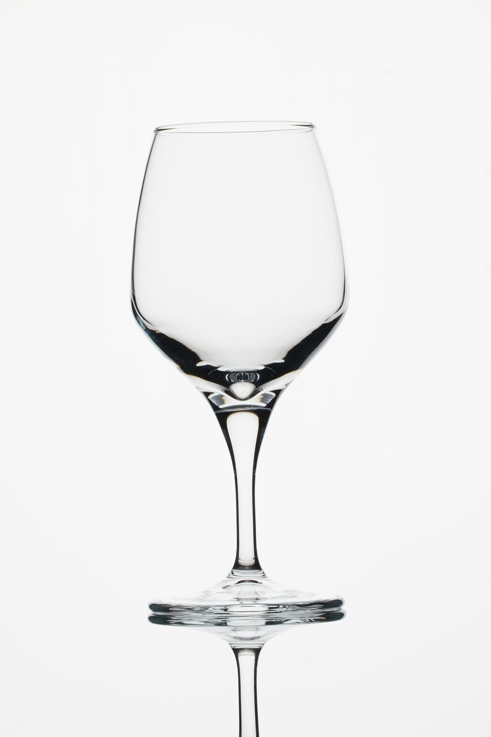 Leeg wijnglas recht van voren tegen een witte achtergrond, met donkere accenten langs de onderkant van de kelk en een spiegeling van de voet en steel in het glanzende tafelblad