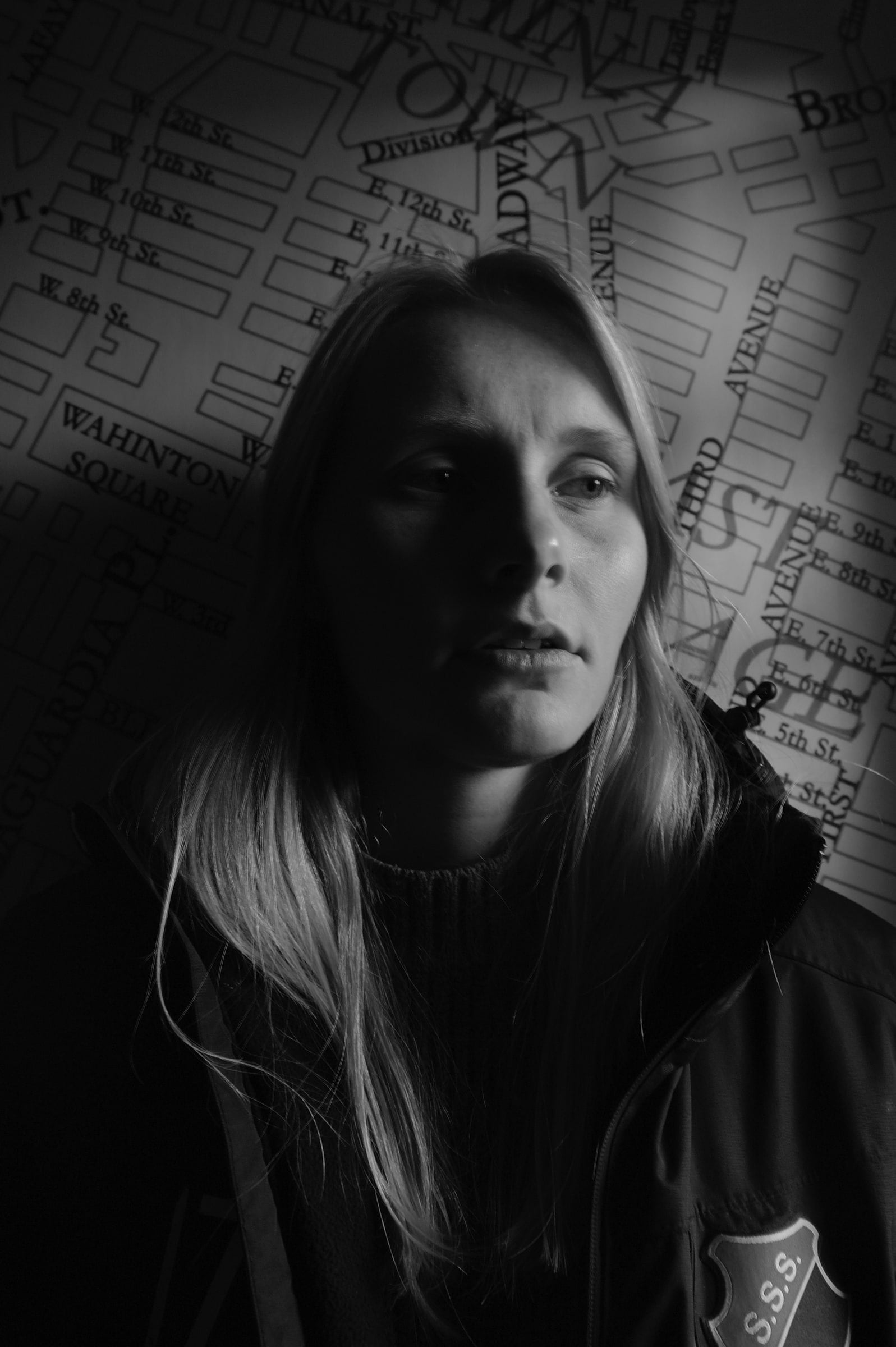Zwart-witportret van een vrouw met lang blond haar in een donkere jas, met een geprojecteerde stadsplattegrond over haar gezicht en de achtergrond