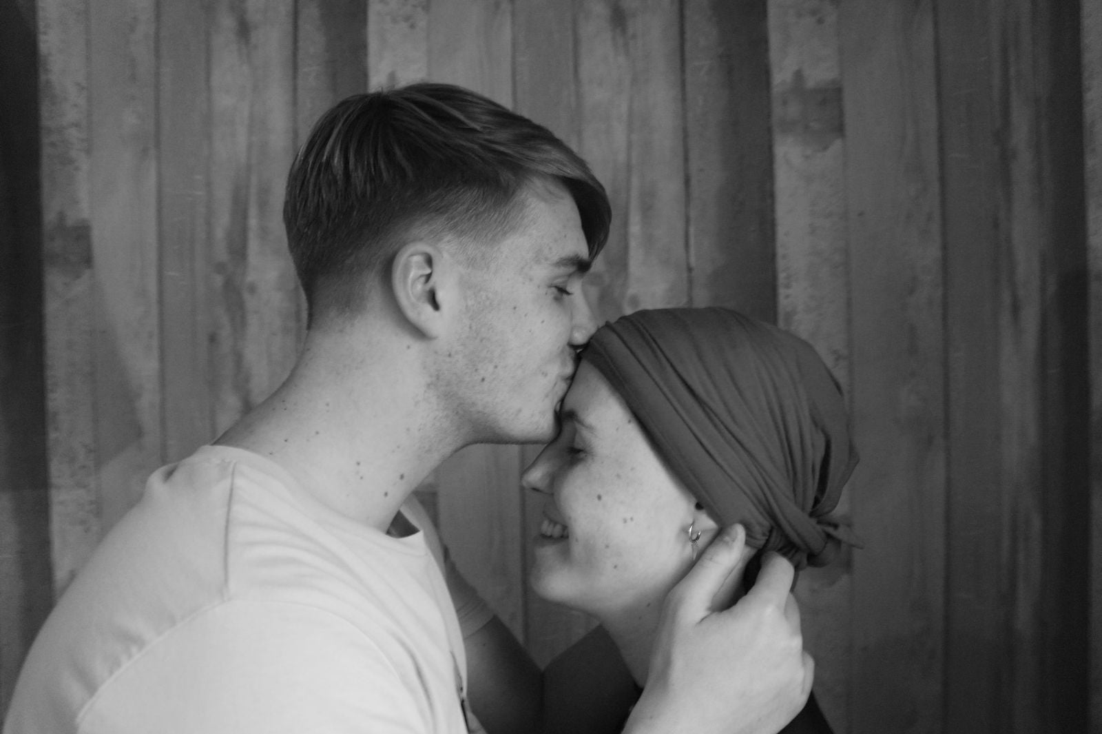 Zwart-wit opname van een jonge man die een vrouw met hoofddoek een kus op het voorhoofd geeft, tegen een houten schuttingwand