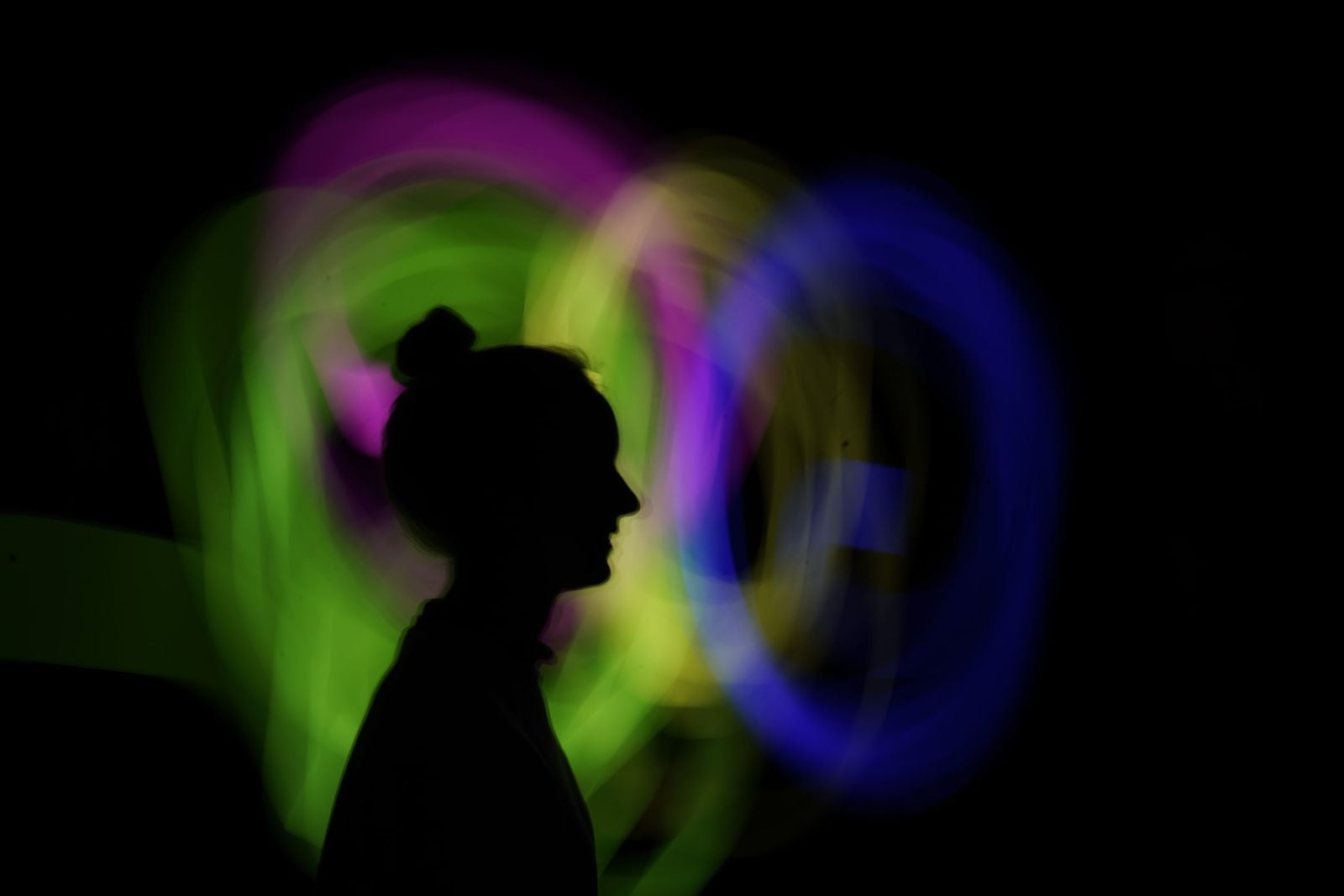 Silhouet van een vrouw met een knot in profiel voor draaiende lichtsporen in groen, roze, geel en blauw tegen een zwarte achtergrond