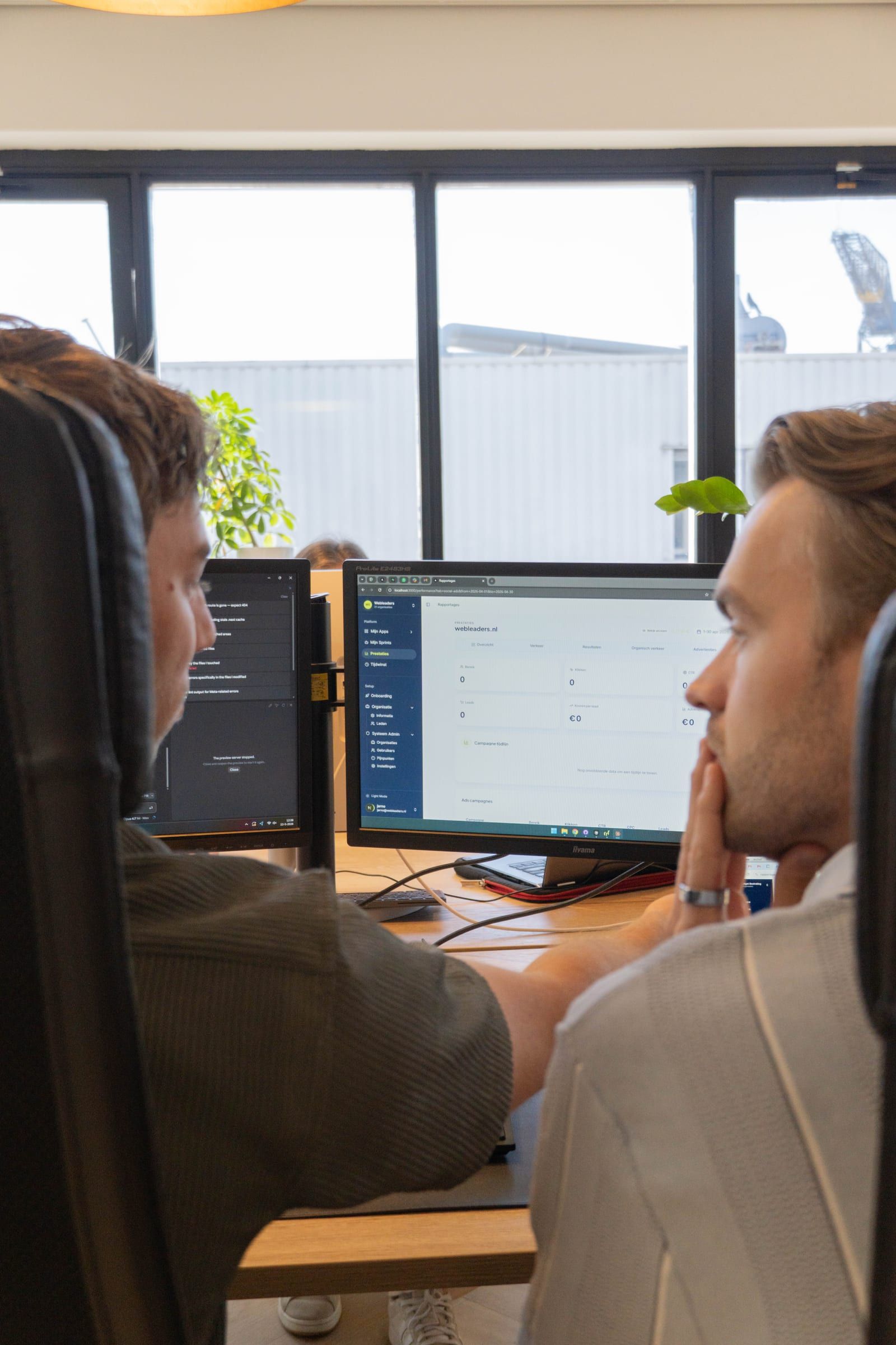 Twee collega's kijken vanaf hun bureaustoelen naar een scherm met een dashboard; op het scherm ernaast staat code.