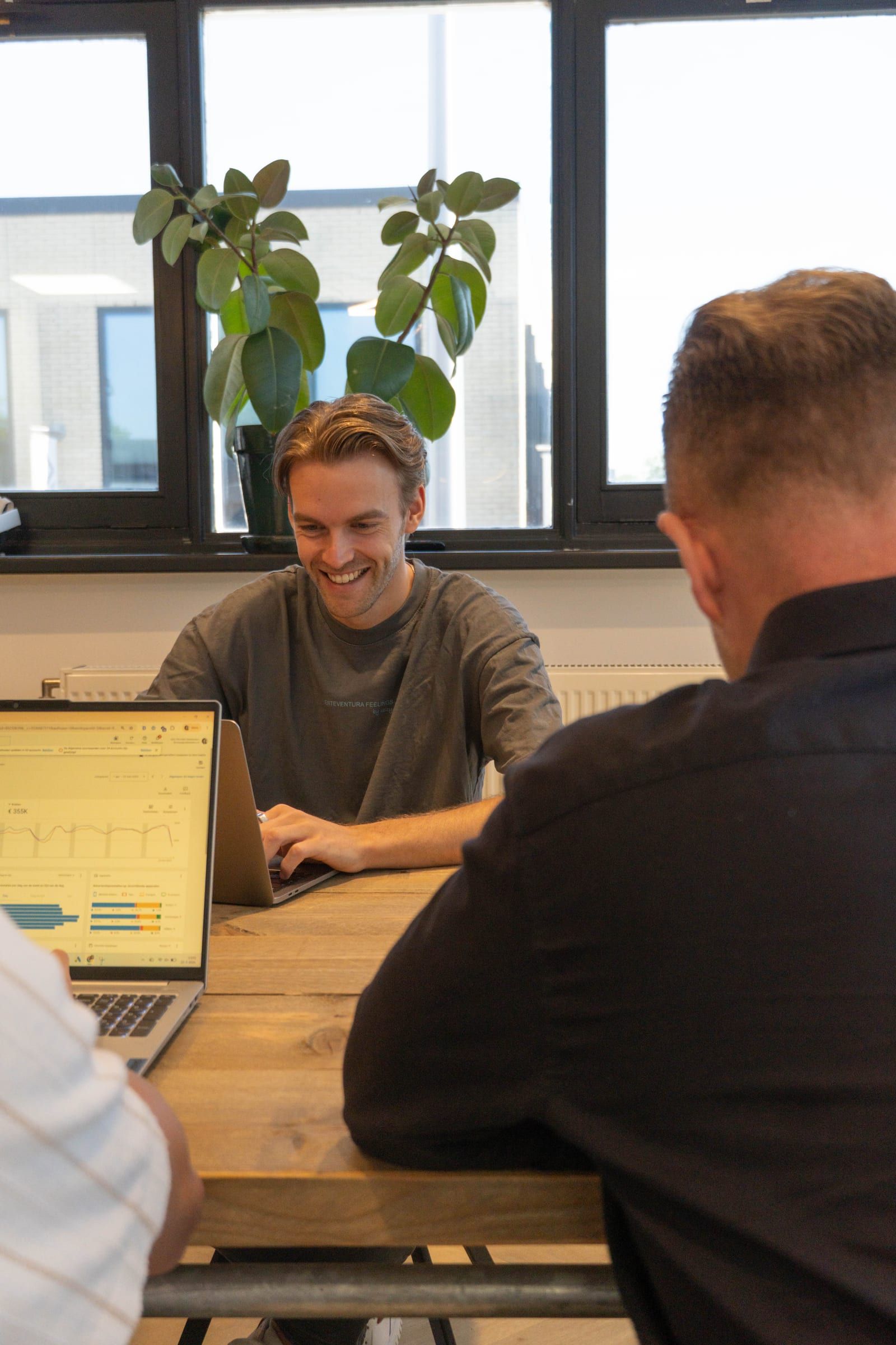Lachende man werkt op een laptop aan een houten vergadertafel, met op de voorgrond twee collega's op de rug gezien.