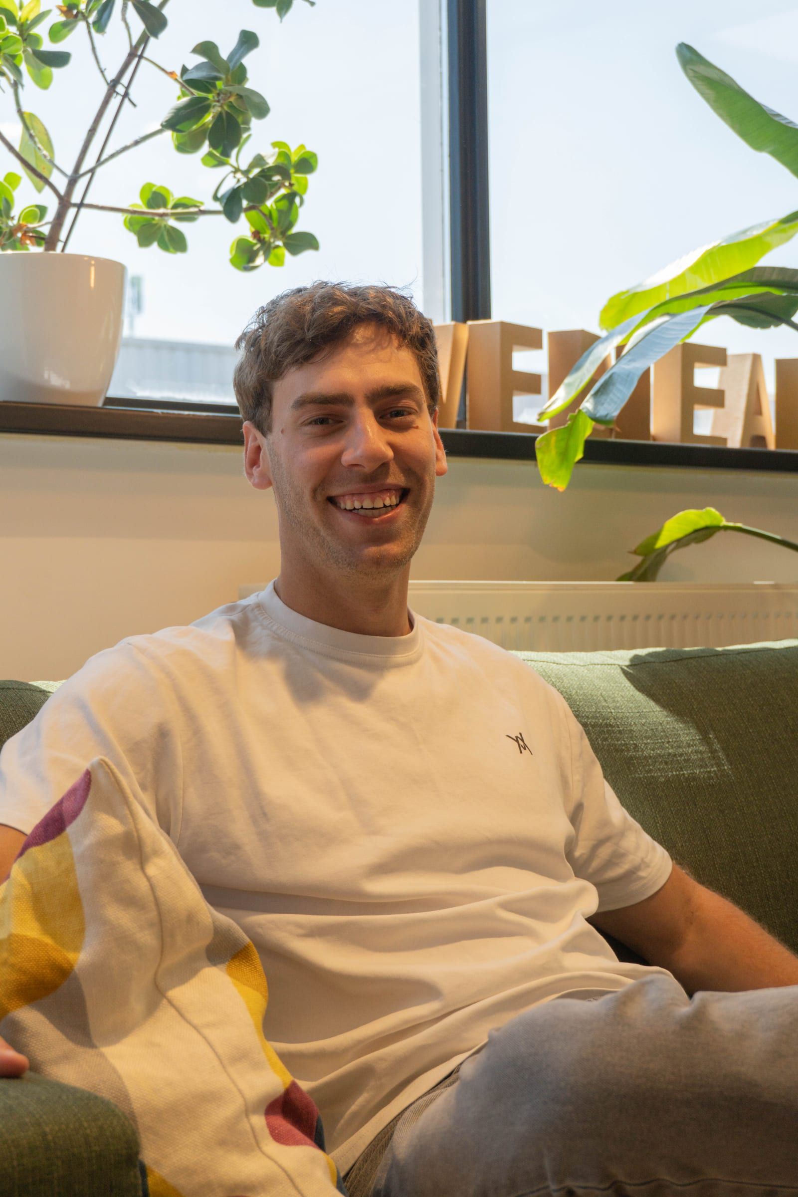 Lachende man in wit T-shirt zit ontspannen op een groene bank, met planten en de Webleaders-letters in de vensterbank achter hem.