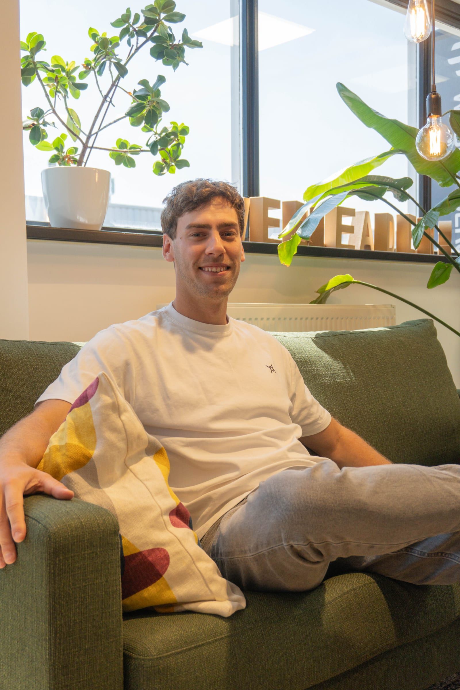 Man zit met een arm over de rugleuning op een groene bank in de loungehoek, omringd door planten en daglicht.