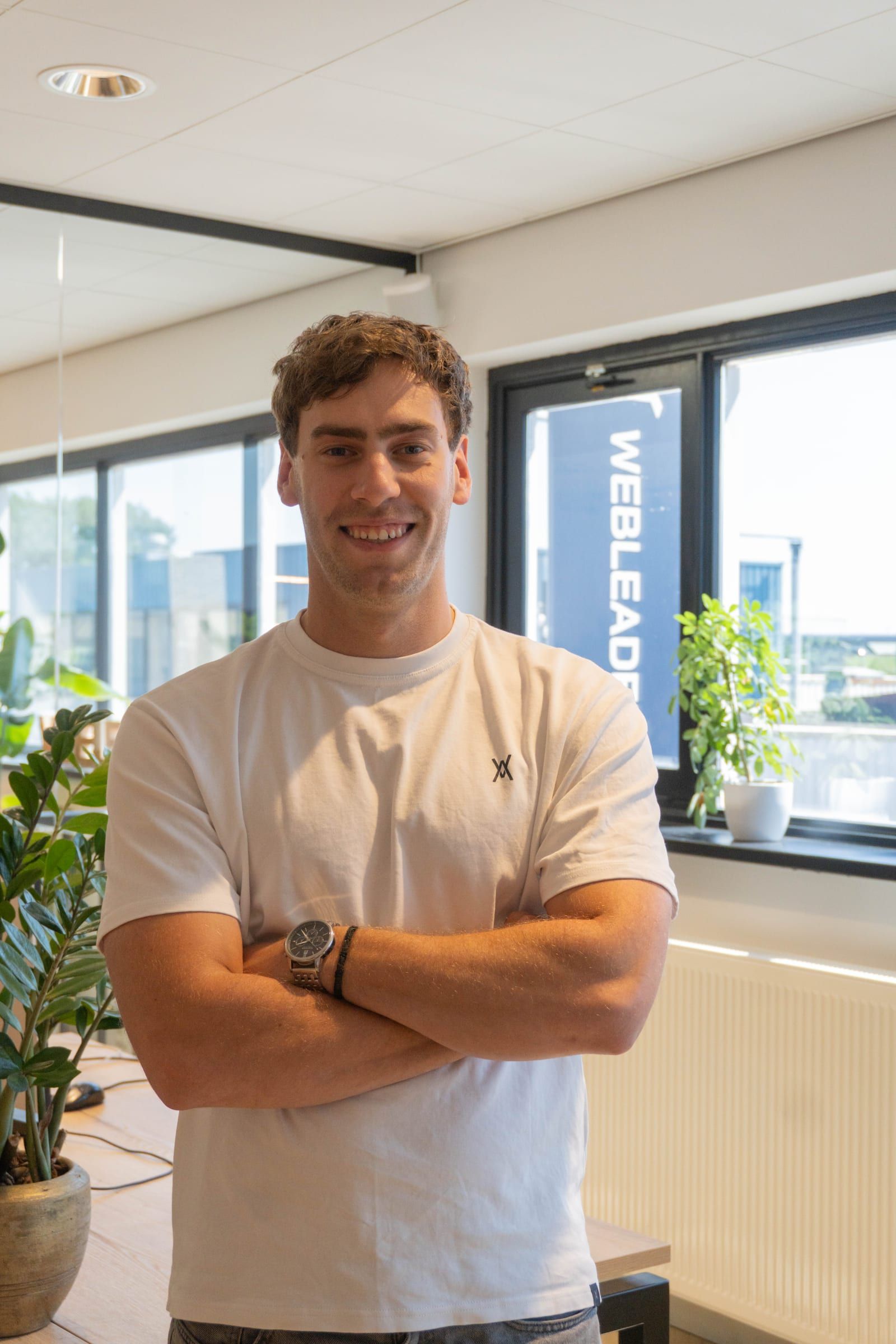 Man met gekruiste armen staat in het kantoor voor een Webleaders-banner en een raam met planten.