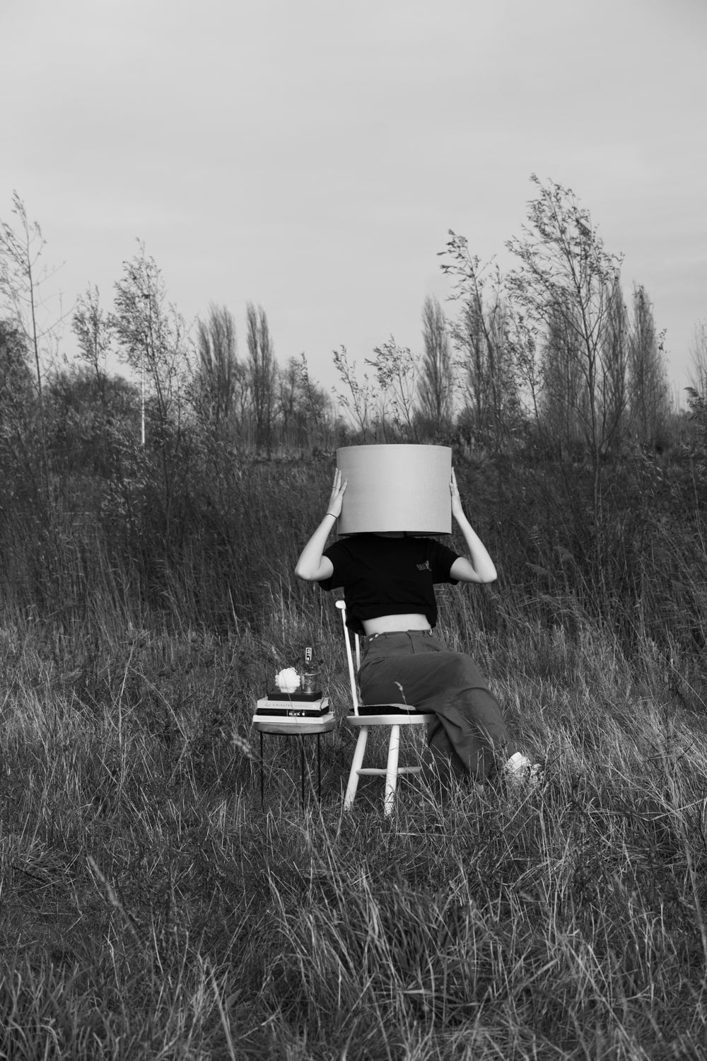 Zwart-wit beeld van een gestalte op een krukje in een grasveld, die met beide handen de lampenkap over het eigen hoofd vasthoudt terwijl de wind door de jonge bomen op de achtergrond waait