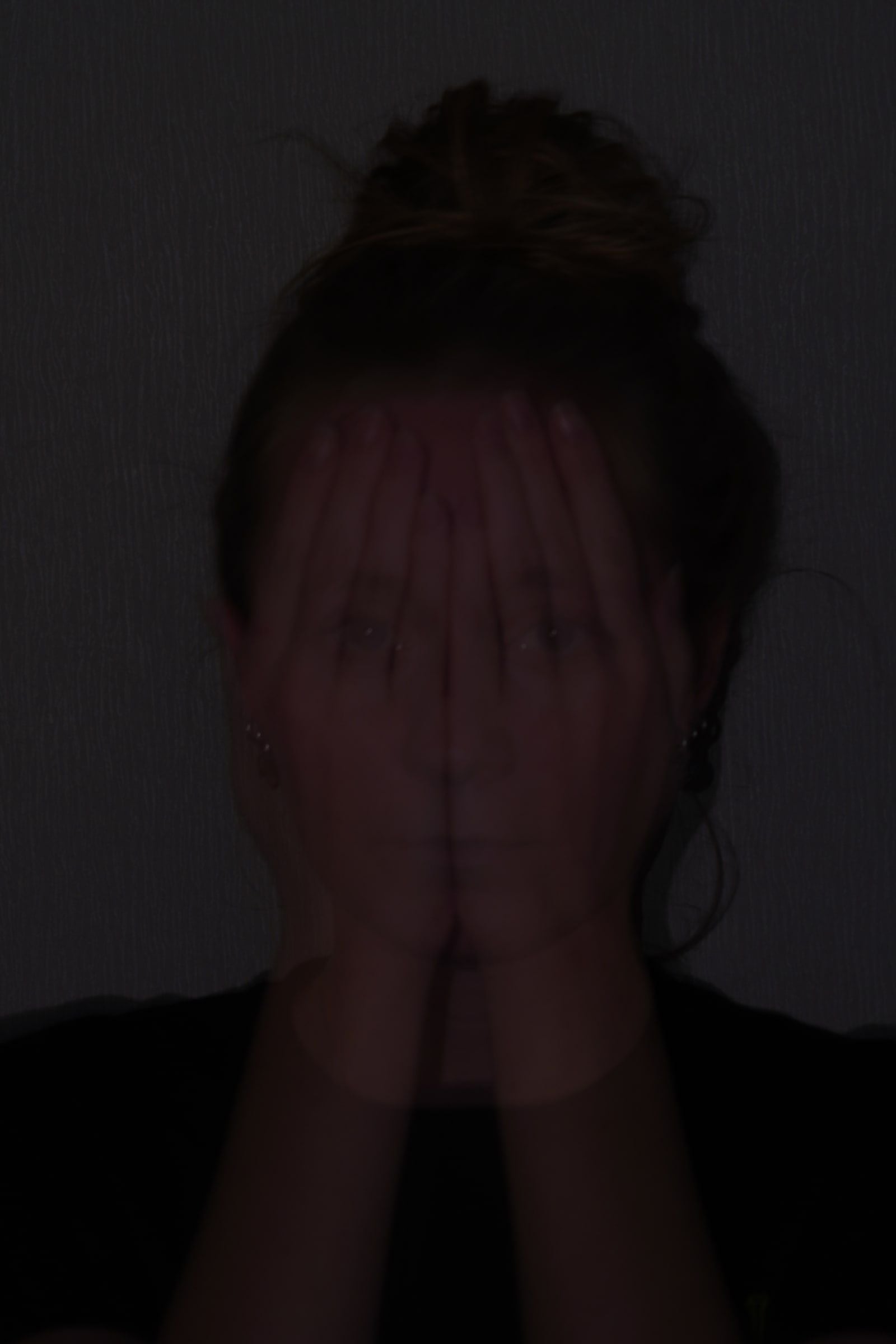 Donker portret waarin twee handen het gezicht bedekken; door de meervoudige belichting schemeren de ogen en de mond door de handen heen, tegen een licht gestructureerde wand